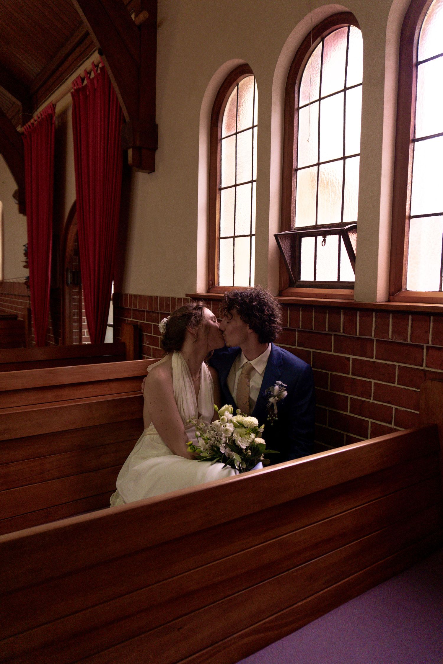 Bride and groom share a romantic kiss in a church pew, bride holding white bouquet, arched windows in background.