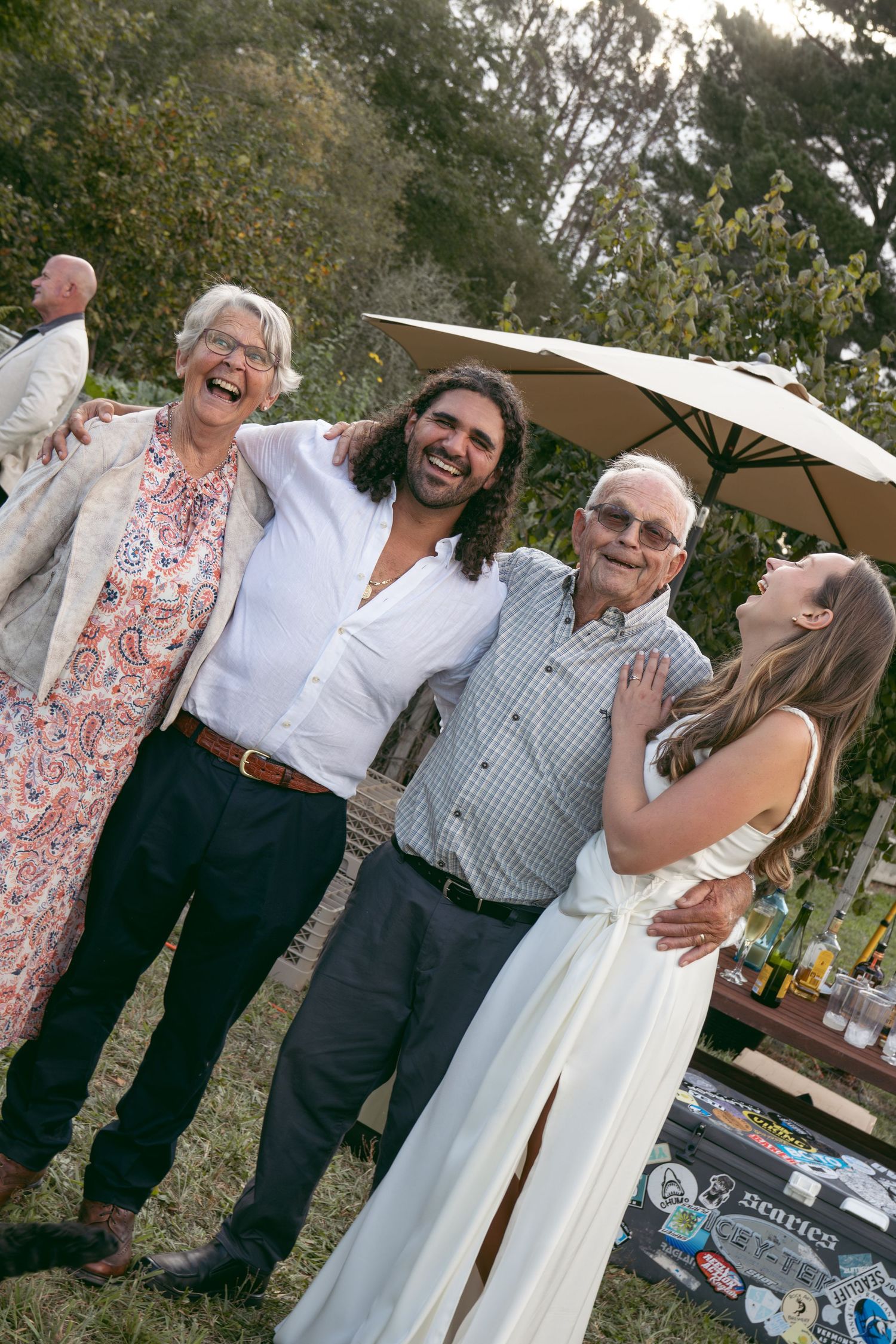Happy family group posing outdoors at a wedding celebration, with bride in white dress and guests laughing together.