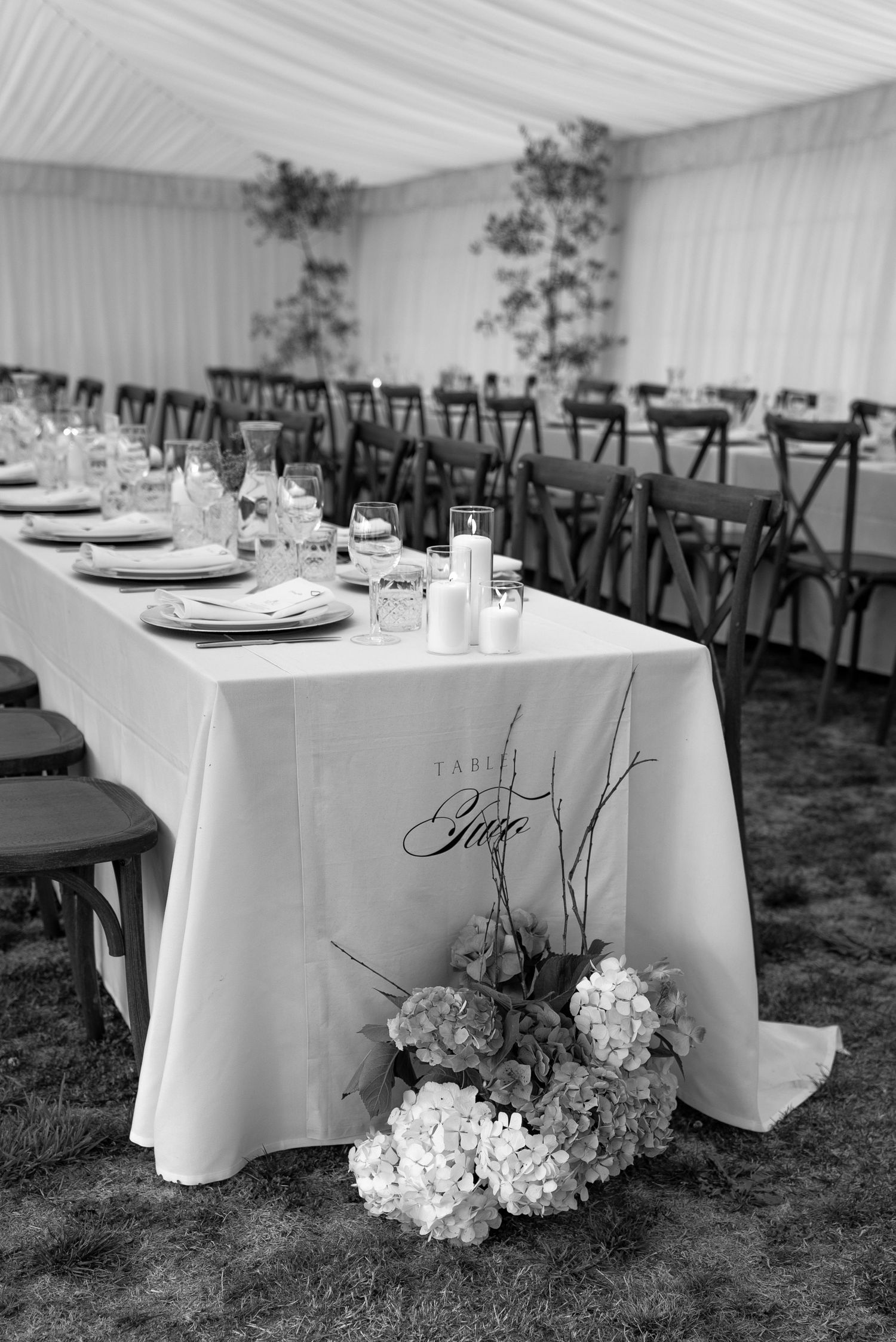 Elegant black and white wedding reception marquee with long tables, floral centerpieces, candles, and hydrangeas on grass floor.