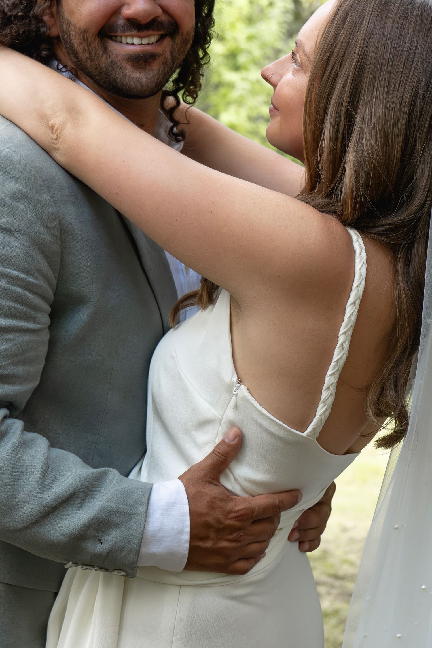 A bride in a white backless gown embraces a smiling groom in a gray suit outdoors.