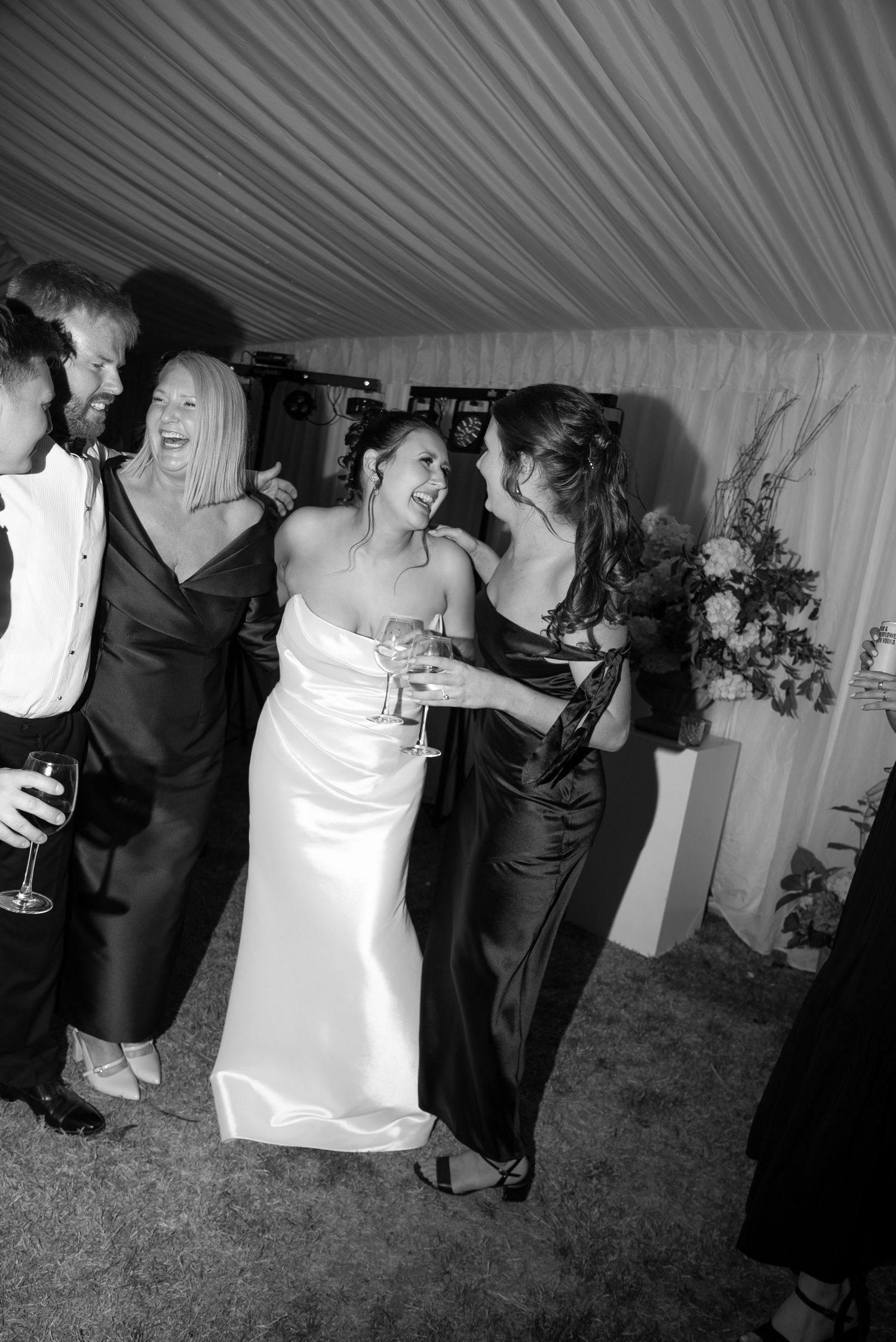 Black and white photo of a bride in white gown laughing with guests in elegant attire at a wedding reception.