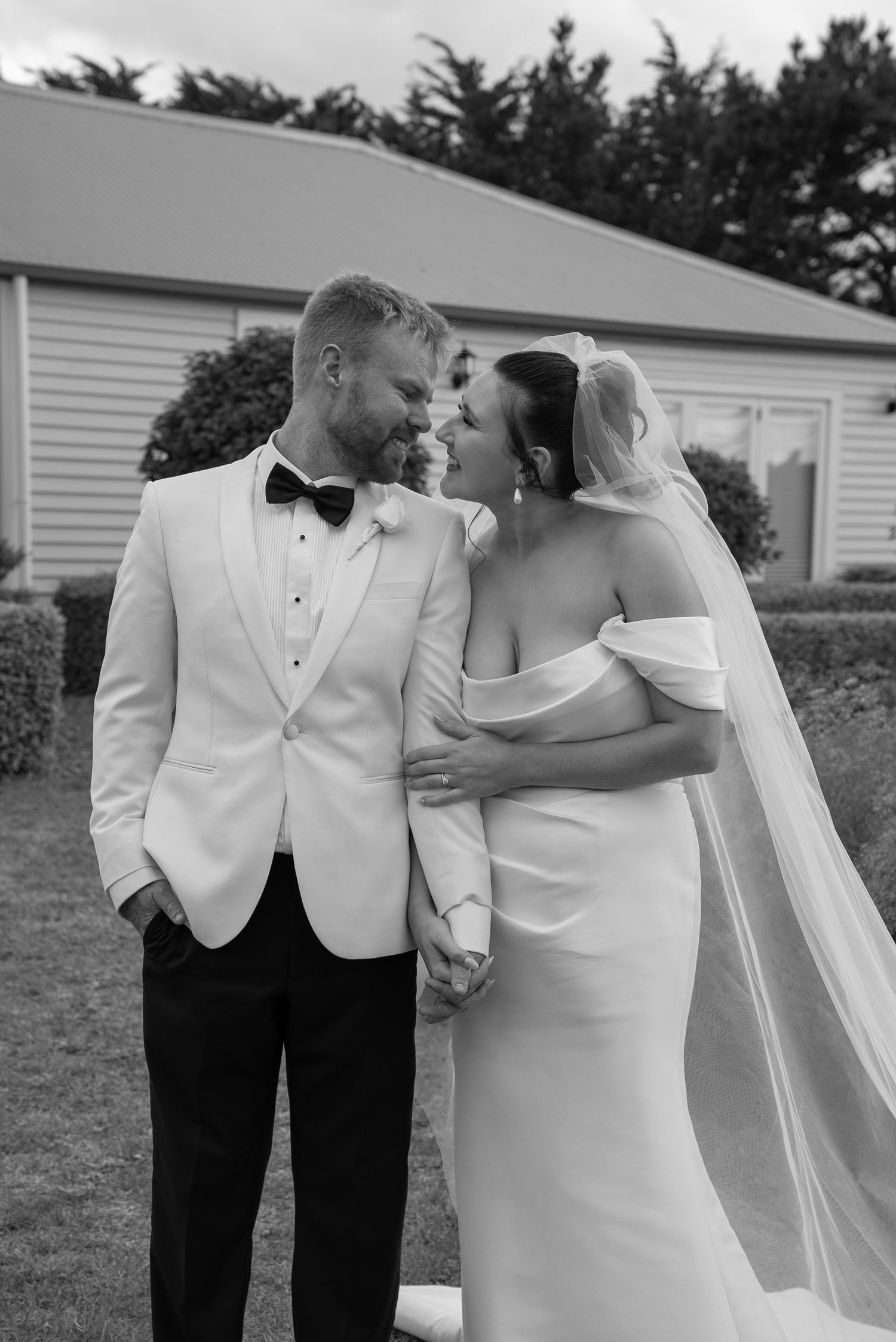 Black and white photo of a bride and groom holding hands and smiling at each other outdoors on their wedding day.
