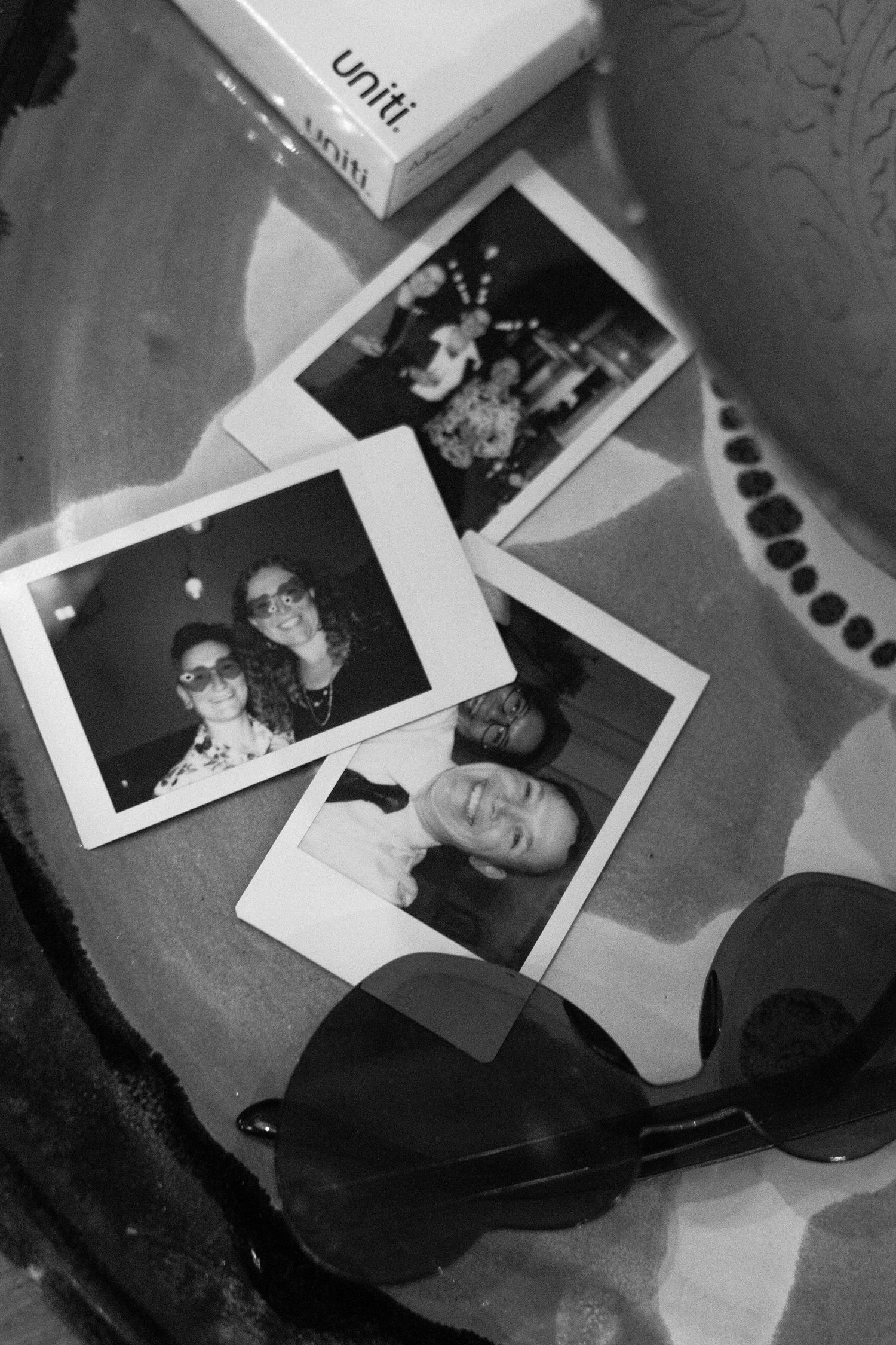 Black and white photo of Polaroid snapshots and sunglasses on a table, with a Uniti box in the background.