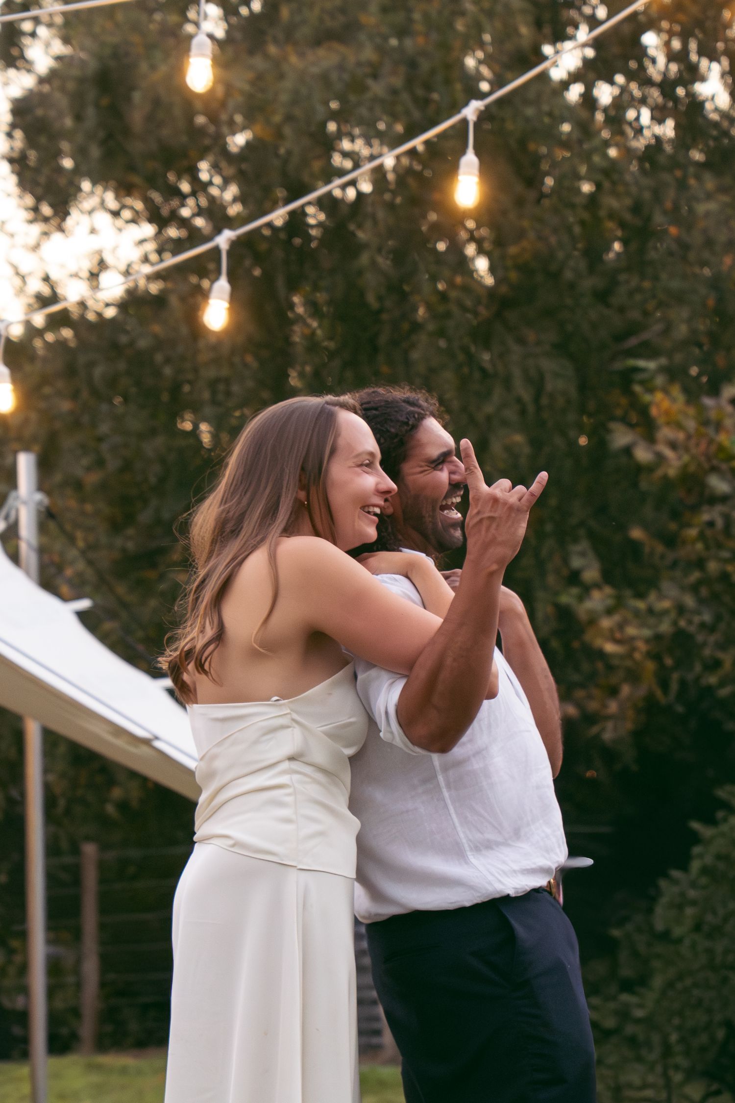 Happy interracial couple dancing outdoors at a garden party with string lights glowing at dusk.