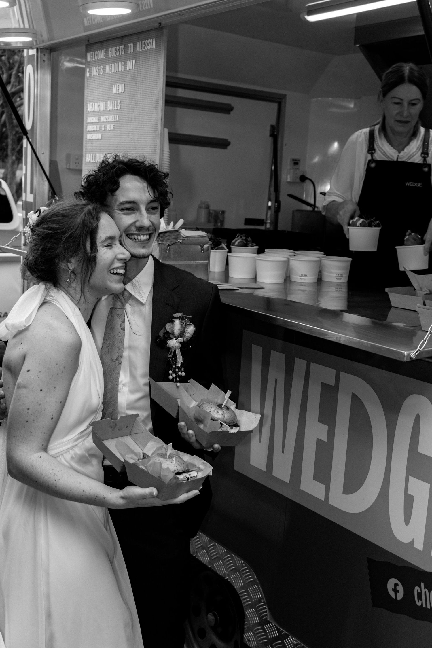 Black and white photo of a laughing bride and groom holding food boxes at a wedding catering truck.