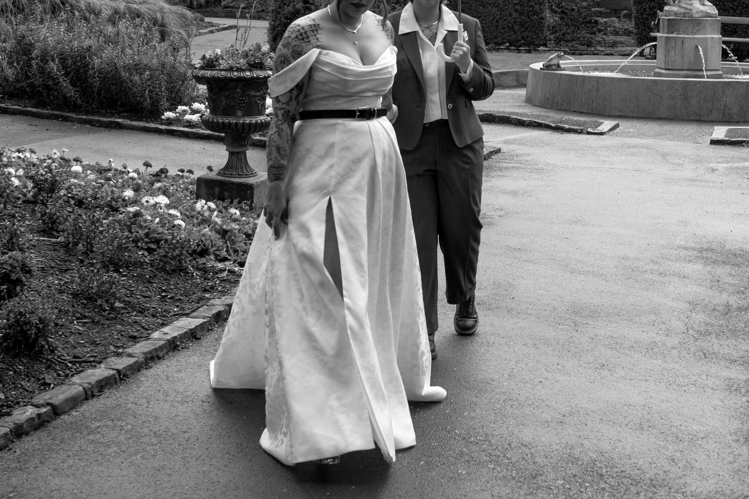 Black and white photo of a bride in a white gown walking with a man in a suit through a garden path.