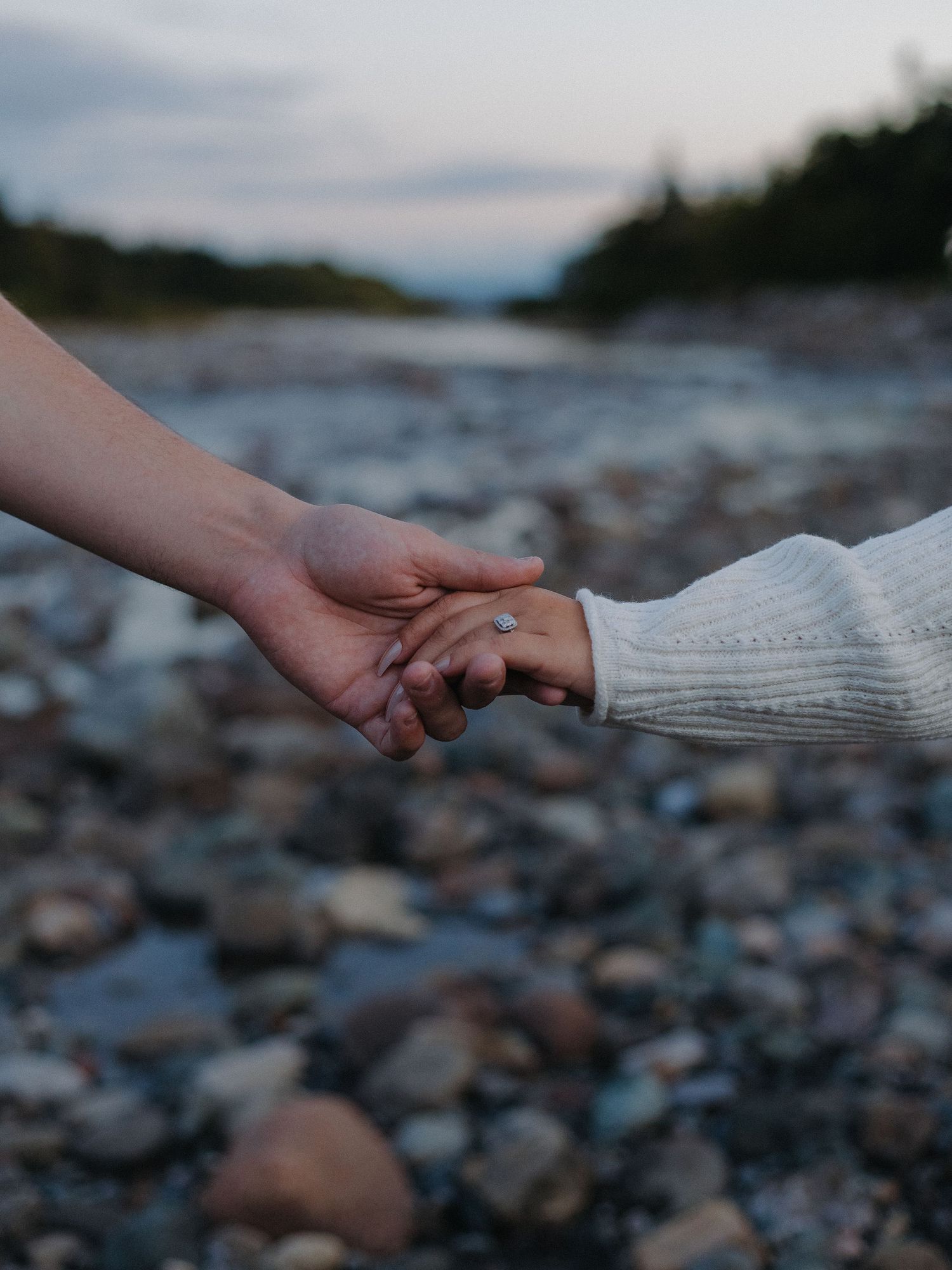 An engaged couple holds hand, showing off her engagement ring, with a sunset in the background.