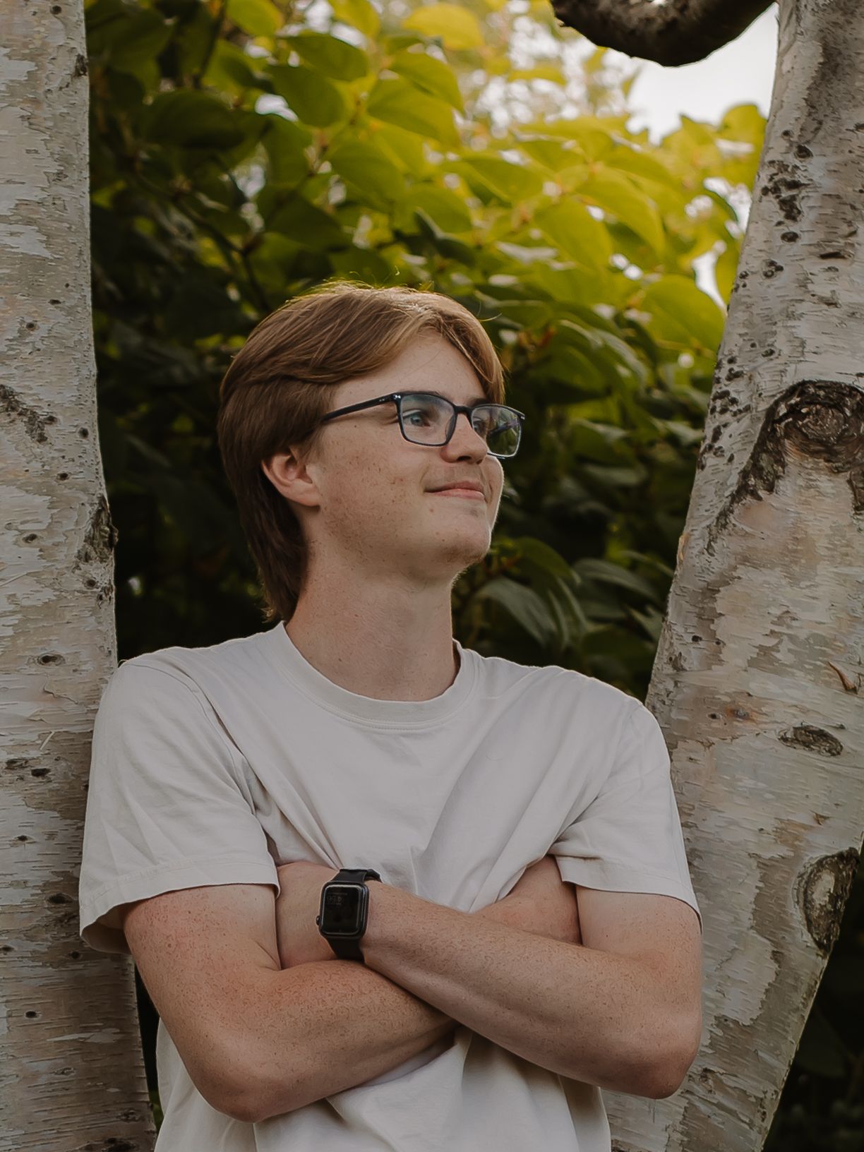 A young teen boy poses against a tree with his arms crossed, looking off into the distance.