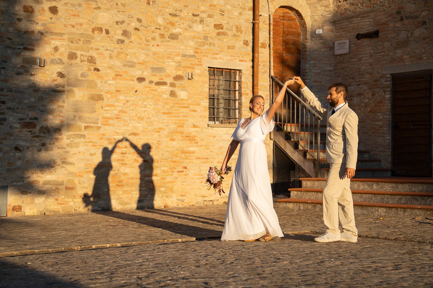 A couple in wedding attire walks along an ancient stone wall in warm evening sunlight.