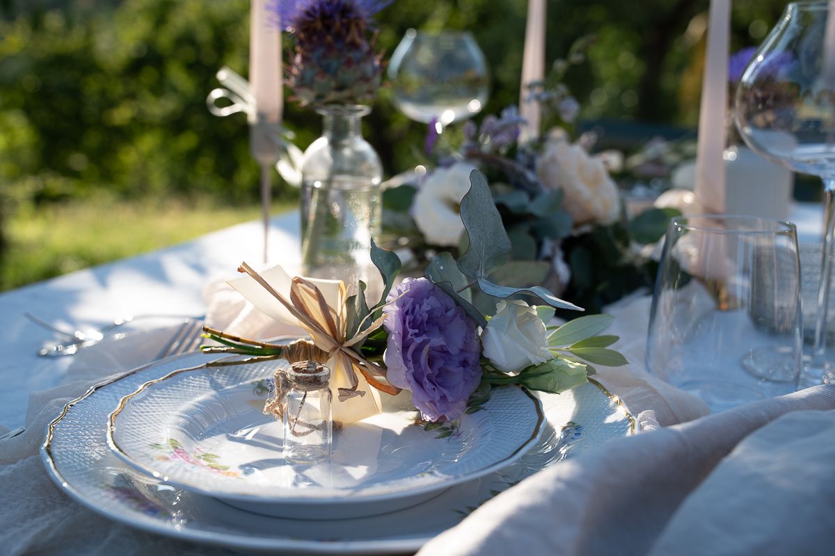 Elegant outdoor wedding table setting with purple roses, white plates, candles, and floral arrangements on a white linen tablecloth.