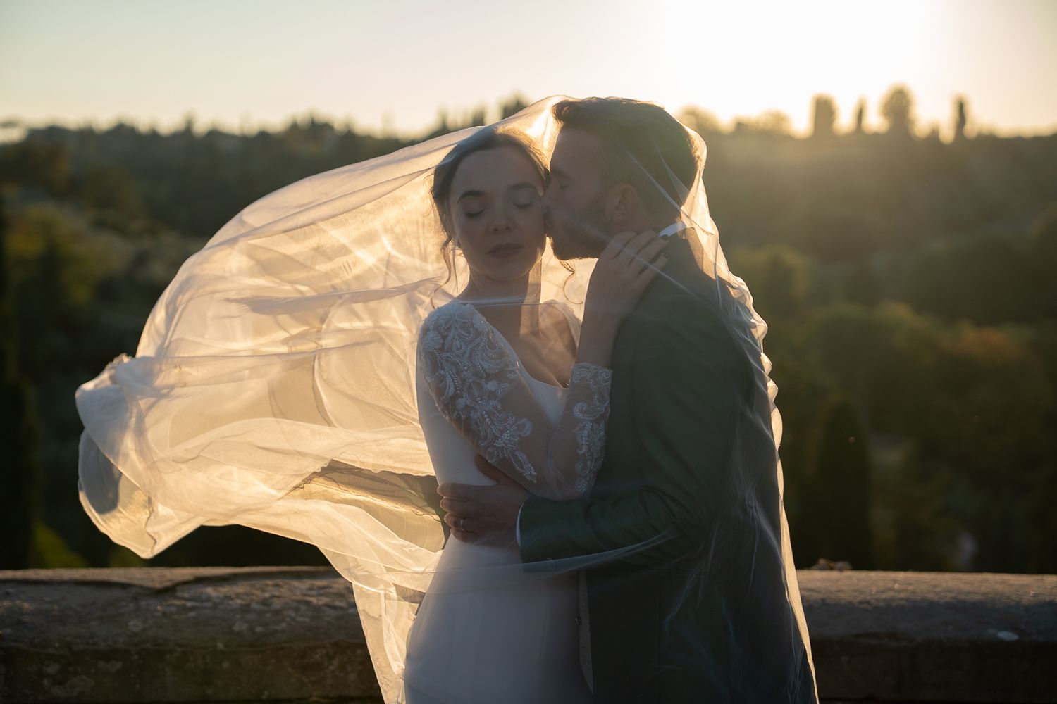 Bride and groom share a romantic kiss at sunset, with veil flowing in the wind against a lush green landscape backdrop.