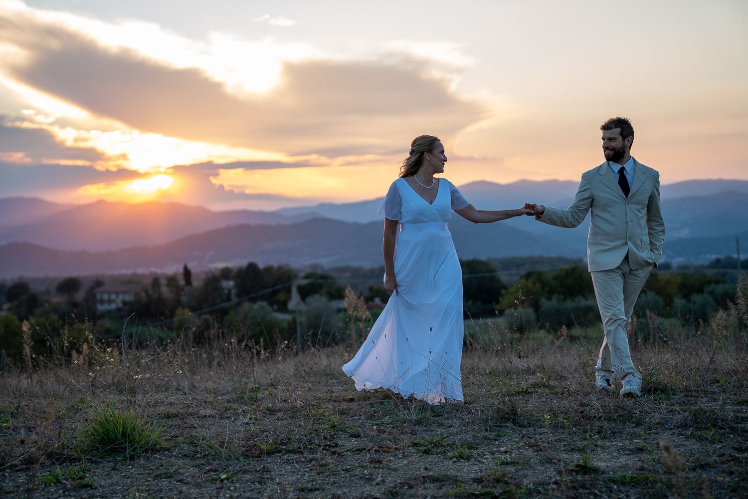 A couple in formal wedding attire dances in a field at sunset with mountains in the background.