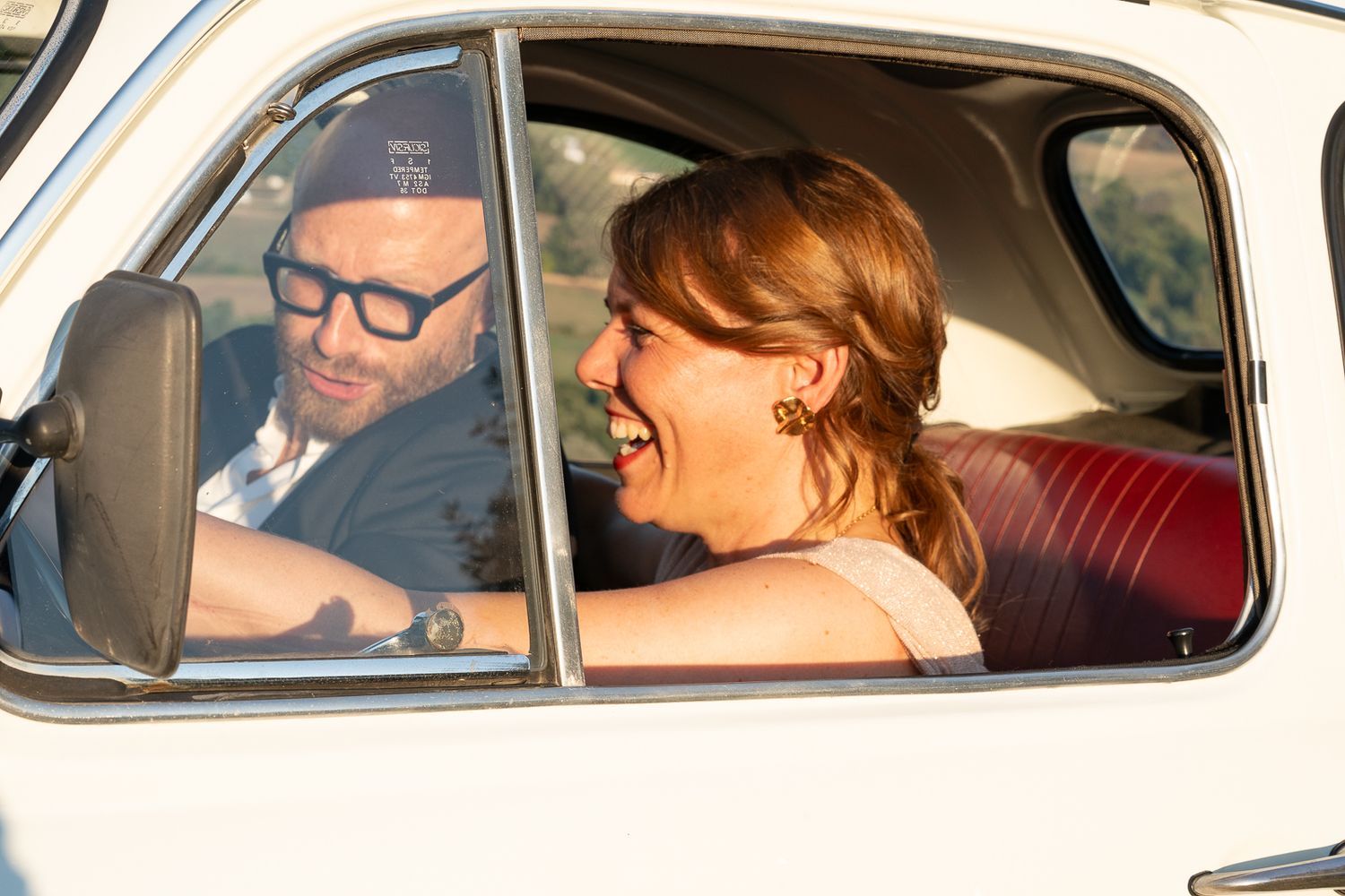 Two occupants sit inside a classic car with red interior visible through the passenger side windows.