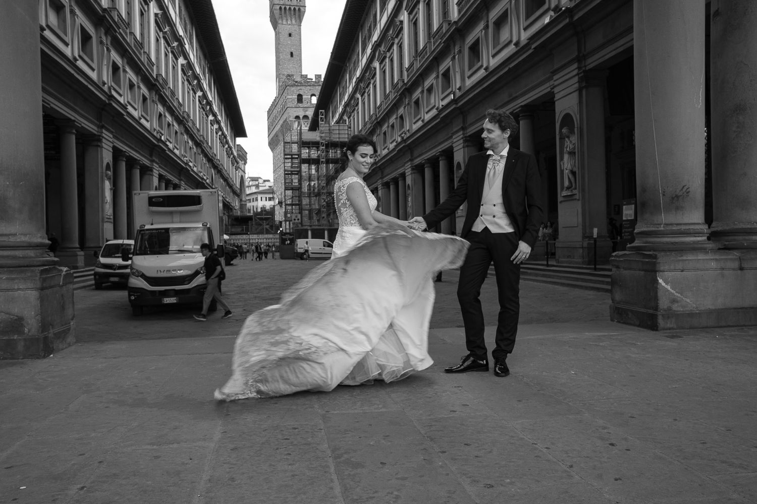 A black and white panoramic sequence shows a bride and groom walking along a historic building's colonnade.
