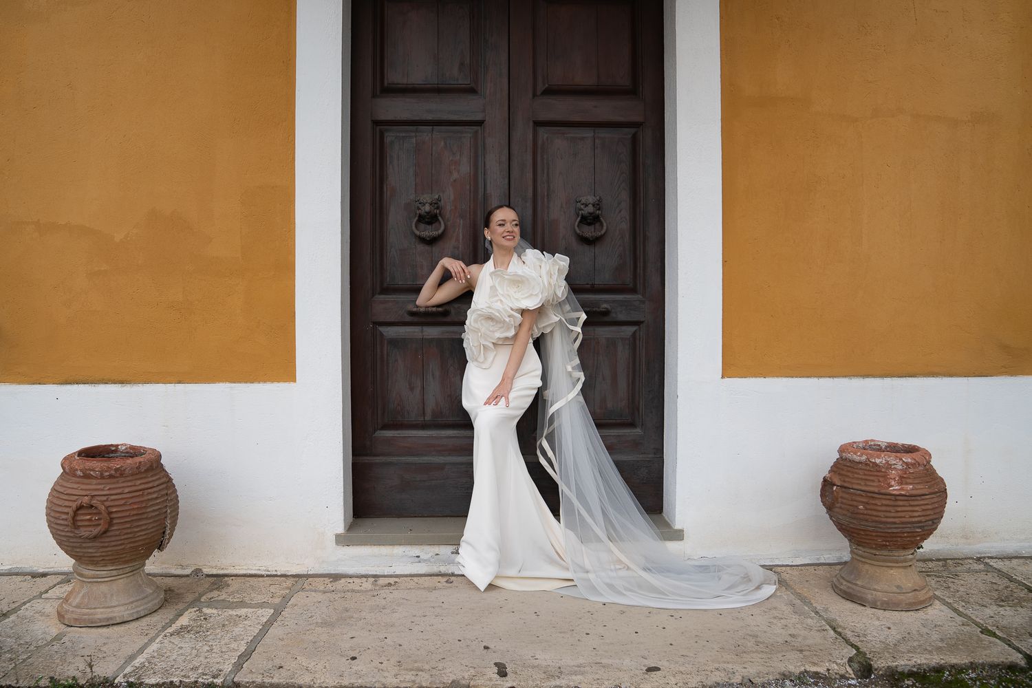A bride in a white dress poses gracefully against a yellow colonial wall with dark wooden doors and terracotta pots.