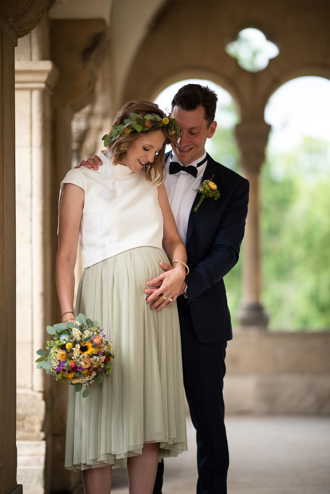 A romantic wedding couple shares an intimate moment under stone arches, with the bride holding a colorful bouquet.