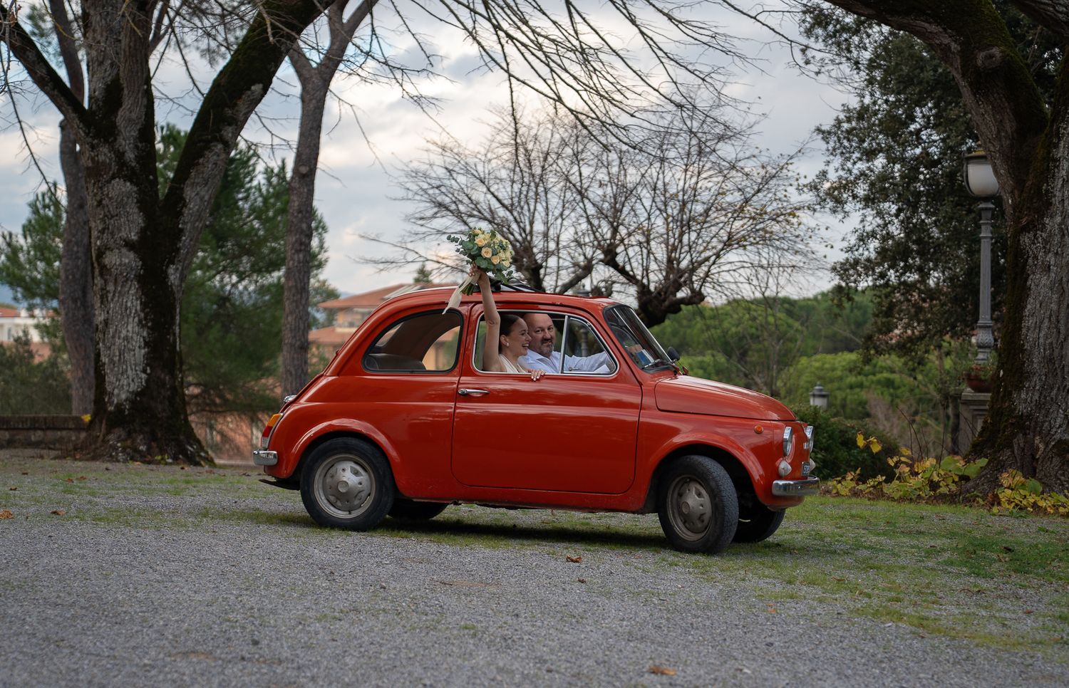 A bright orange vintage Fiat 500 car parked on a gravel path near trees at sunset.