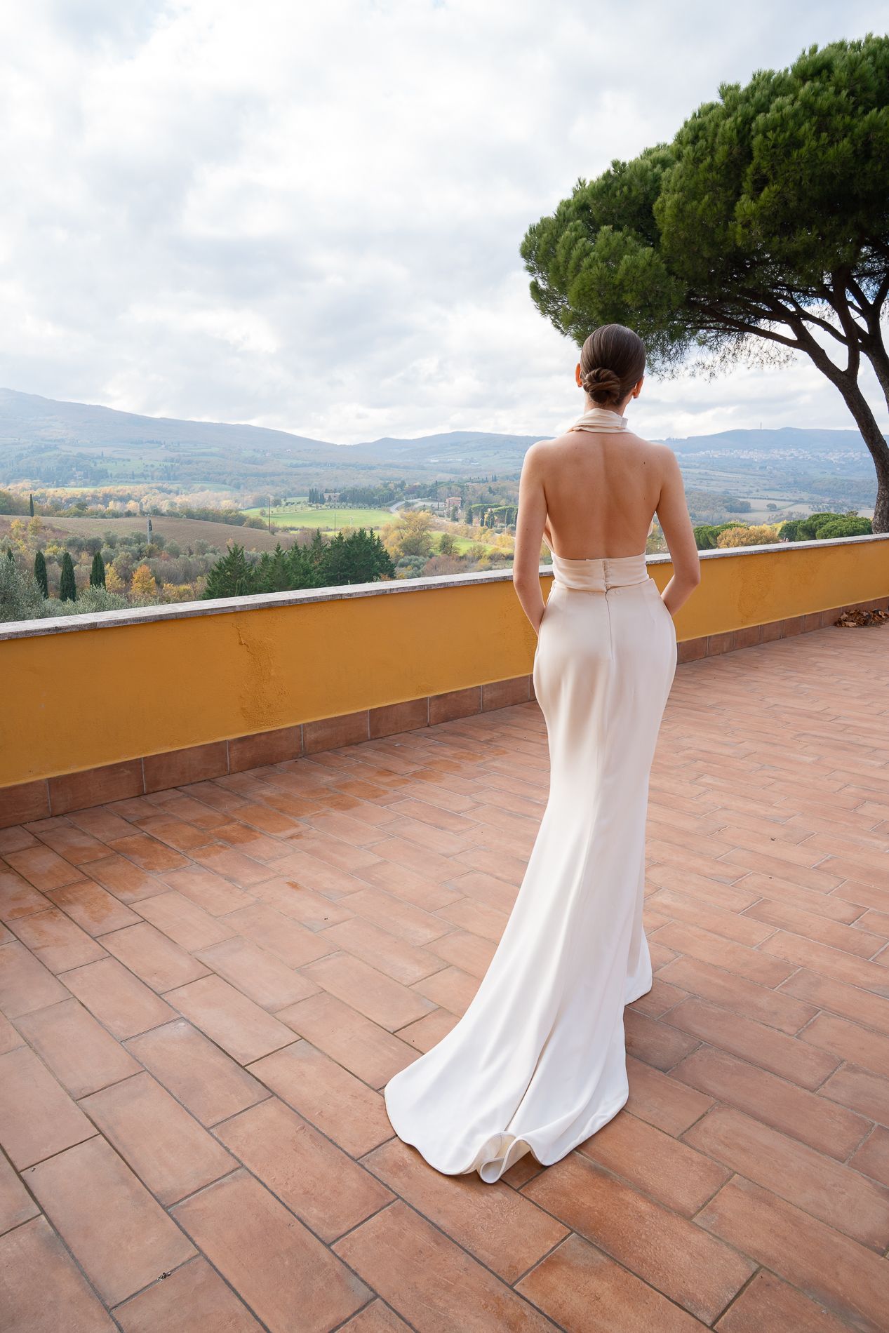 Elegant white wedding dress with open back design shown on terrace overlooking Tuscan countryside landscape.