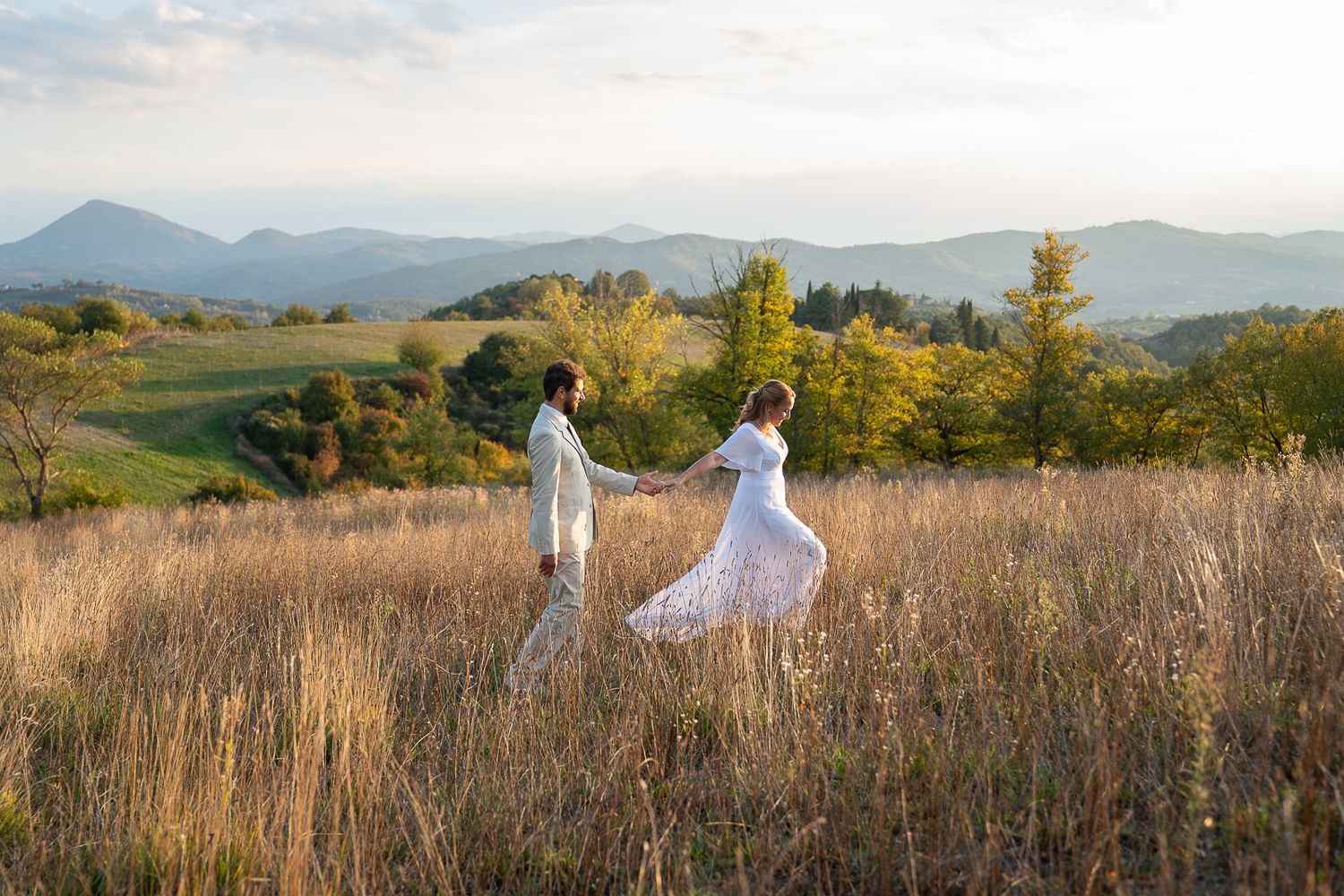 Two people in white dresses walk through a golden field at sunset with mountains in the distance.
