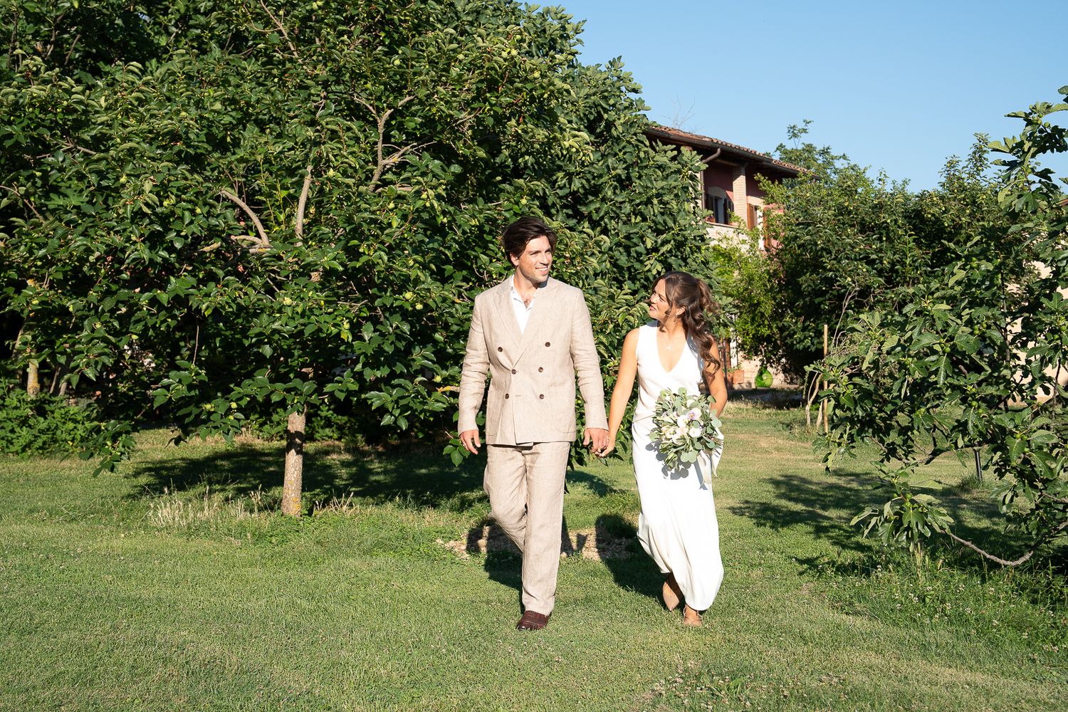 A couple in wedding attire walks hand-in-hand through a lush green garden, on their wedding day at Casale San Cristoforo in Umbria, Italy.