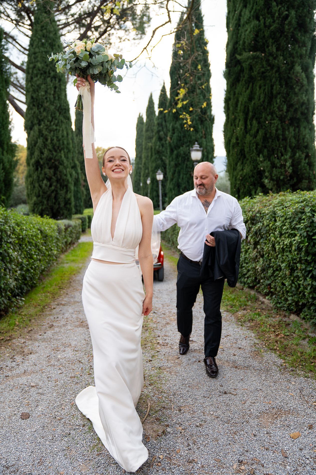 A playful couple in wedding attire runs and jumps along a cypress tree-lined gravel path in Italy.