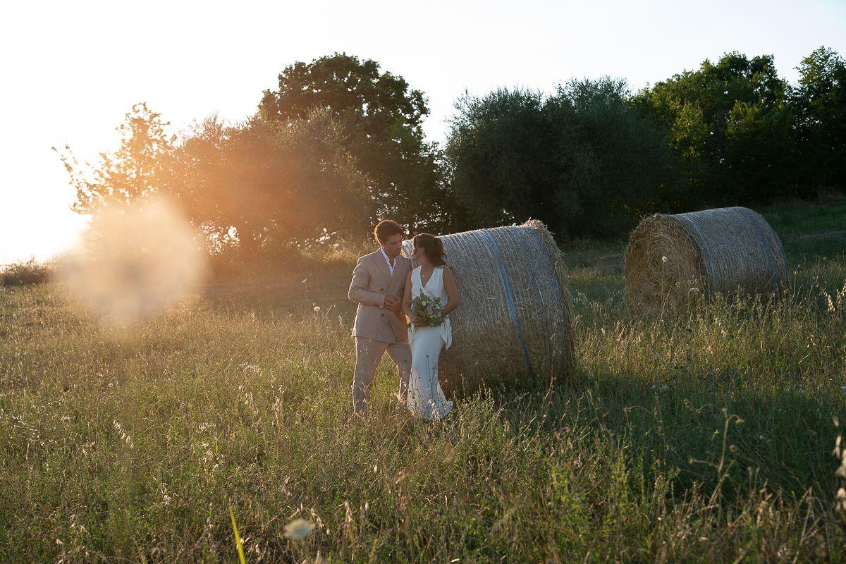 Couple walking in golden sunlit field with hay bales at sunset, romantic outdoor wedding photography in Umbria.
