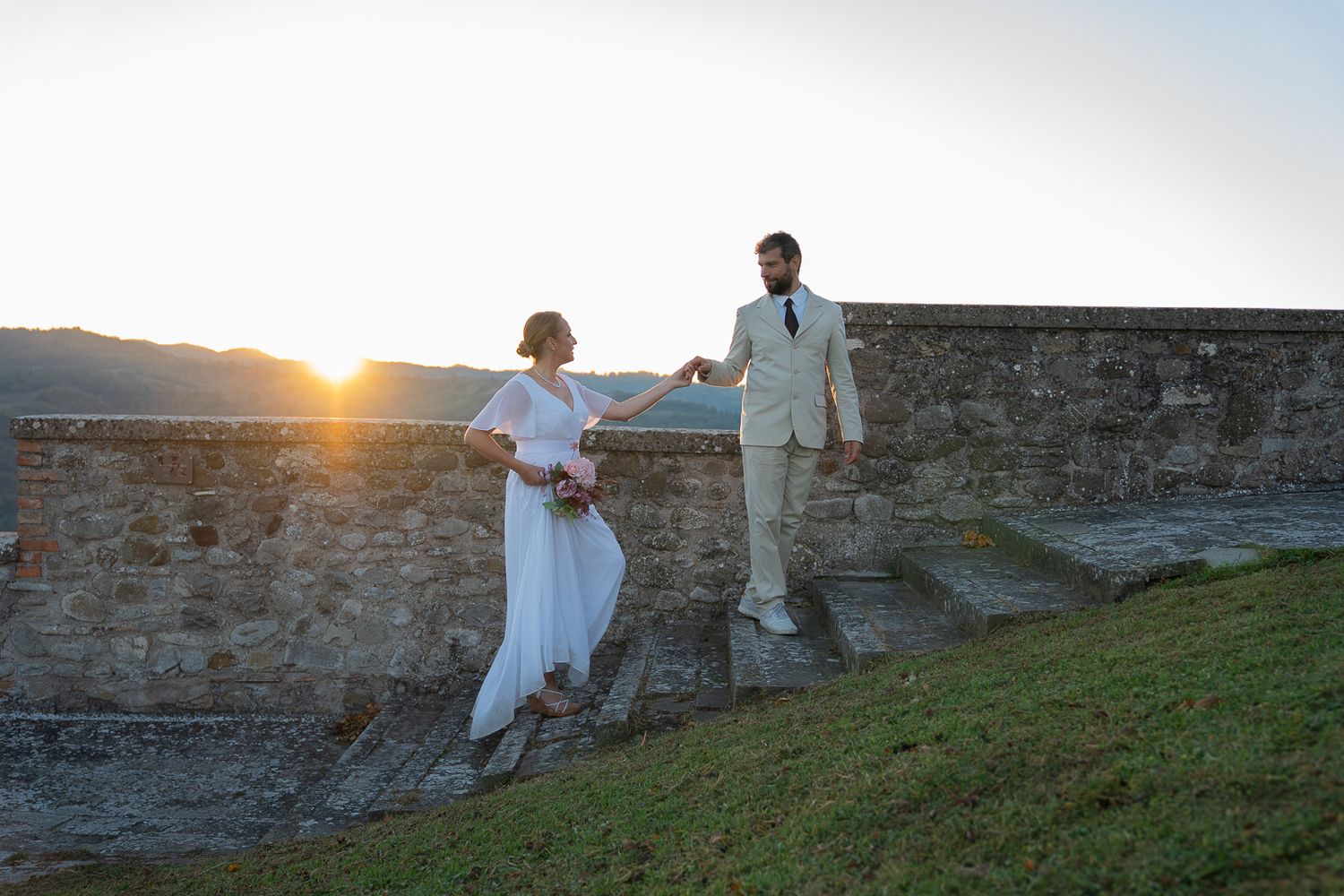 A couple in white outfits stands together on stone fortification walls at sunset creating a romantic silhouette in Montone, Umbria, Italy