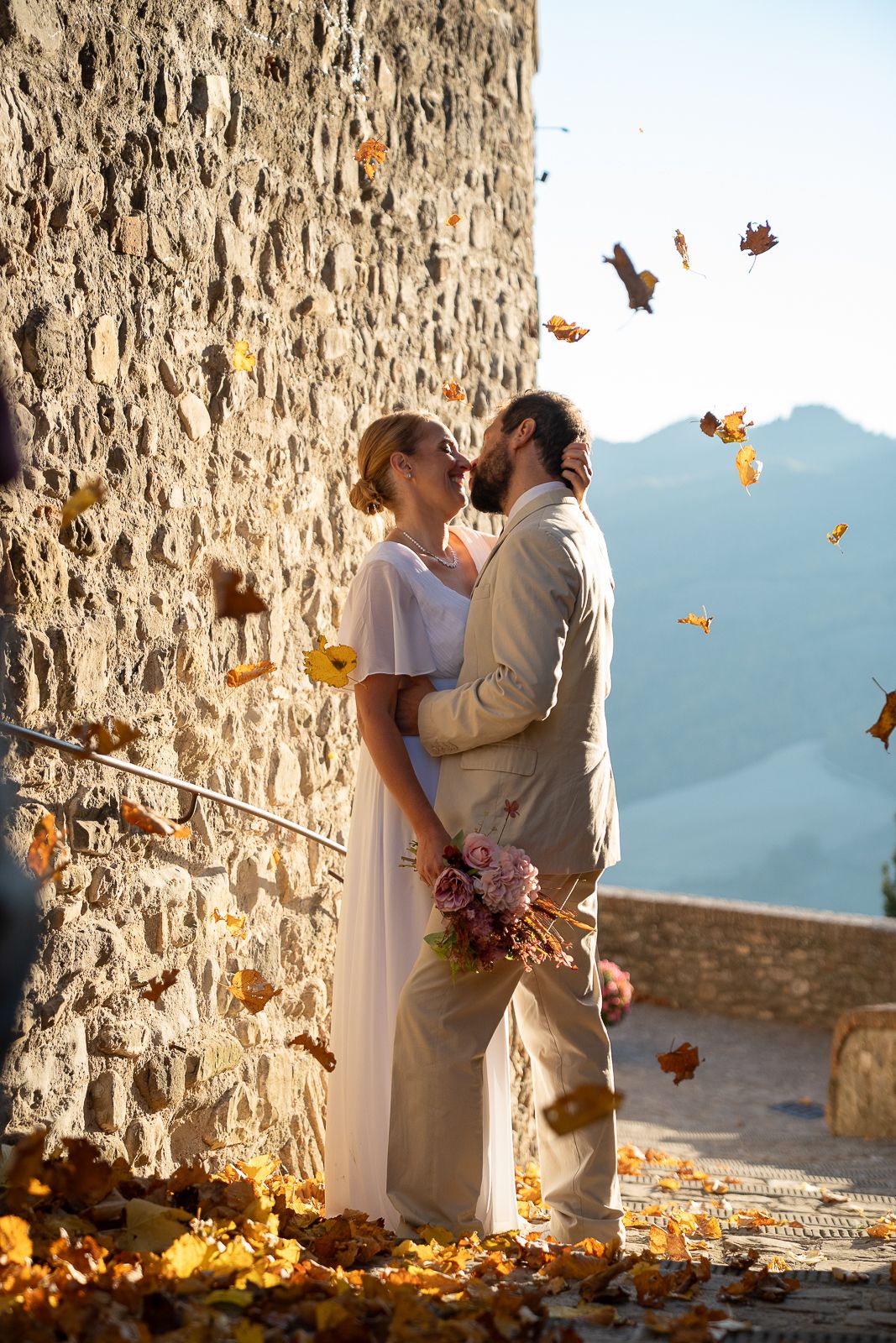 A romantic autumn wedding moment captured against a stone wall with mountains and falling leaves in the background In Umbria, Italy