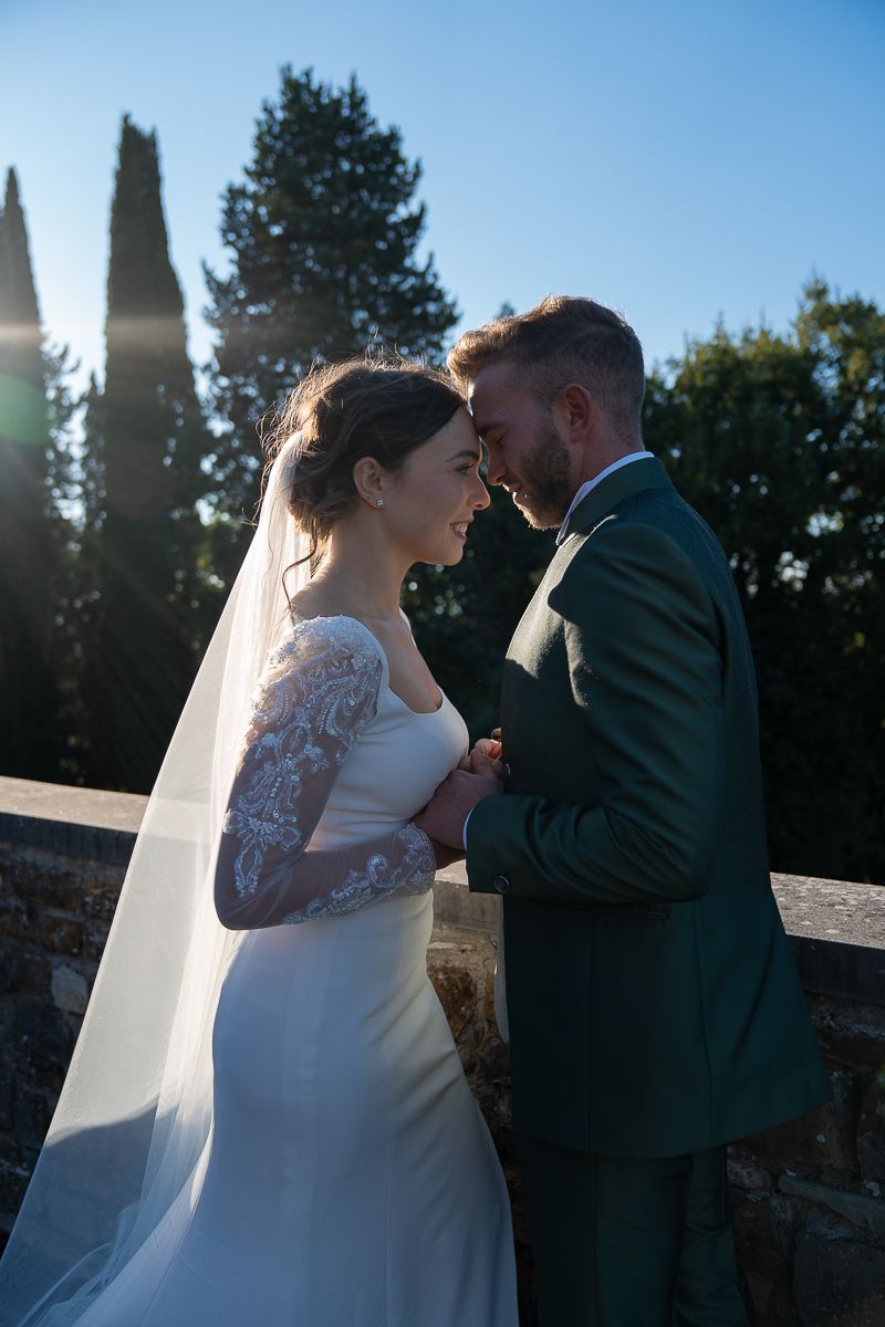 Newlywed couple shares romantic moments at sunset against stone wall backdrop in wedding attire.