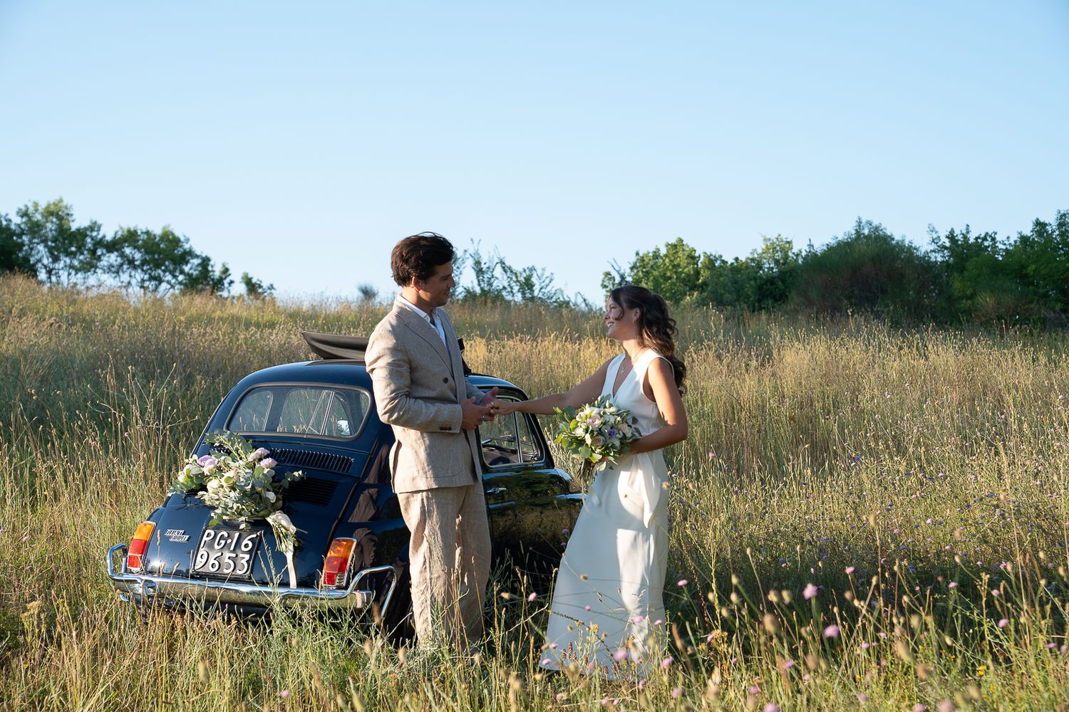 Bride and groom standing by a vintage blue car in a golden meadow on their wedding day.