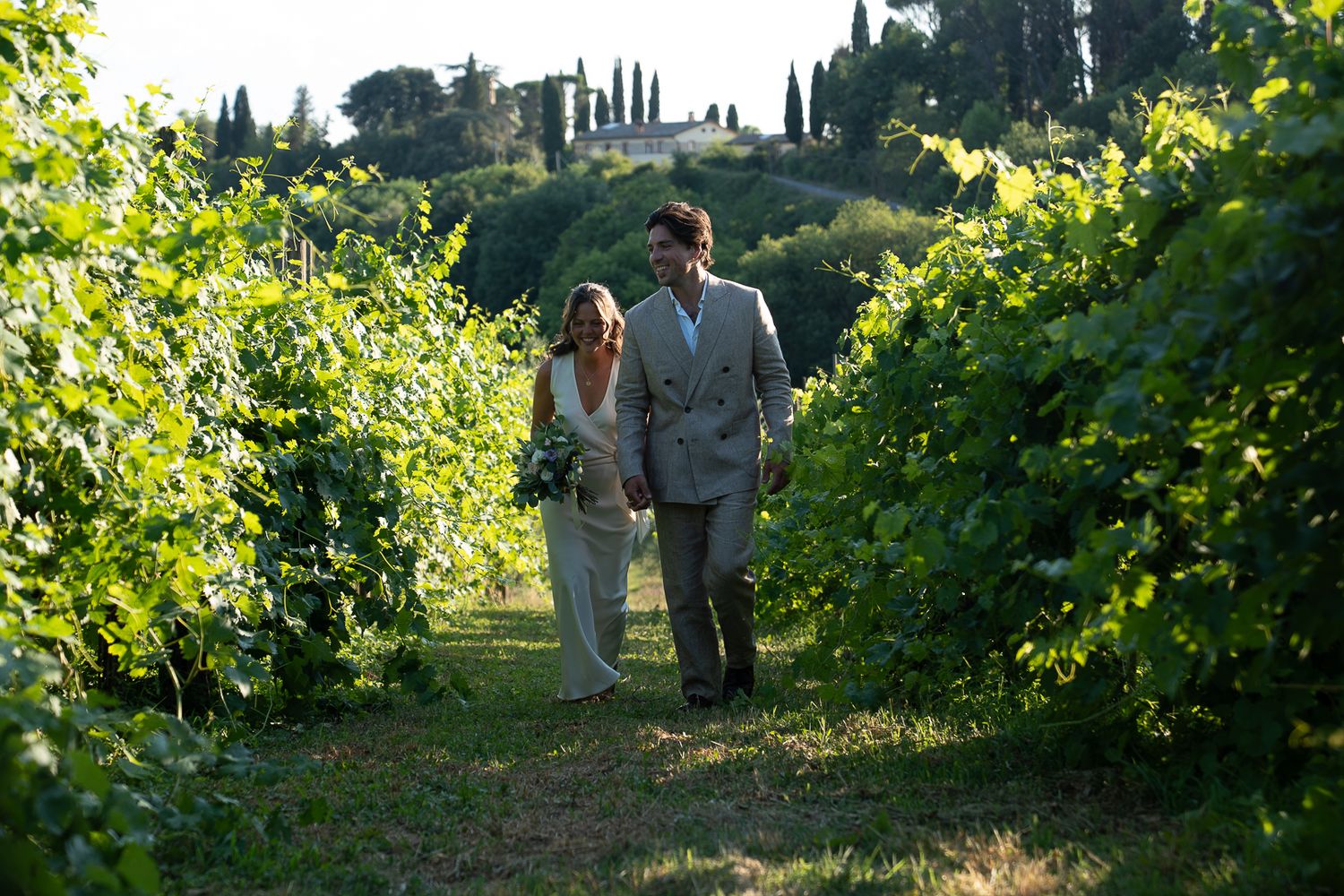 A couple walks hand-in-hand through lush vineyard rows at a Tuscan estate wedding, surrounded by green vines.