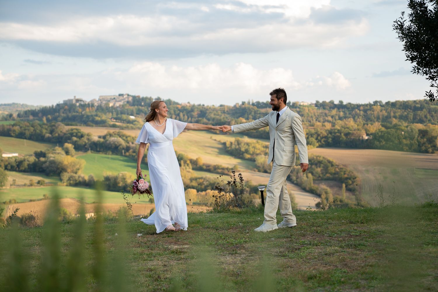 Couple in white outfits dancing together on a hilltop overlooking a scenic countryside landscape at sunset.