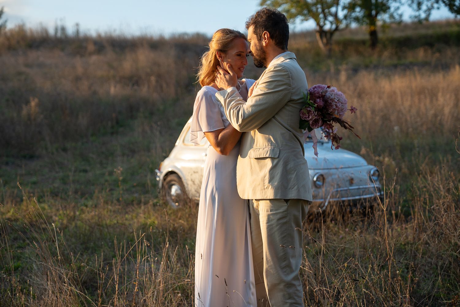 A romantic couple in wedding attire embrace in a golden field at sunset with a vintage white Fiat 500 car in the background.