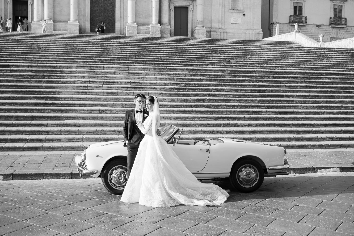 Black and white wedding photo of bride and groom embracing by a vintage convertible car on grand stone steps.