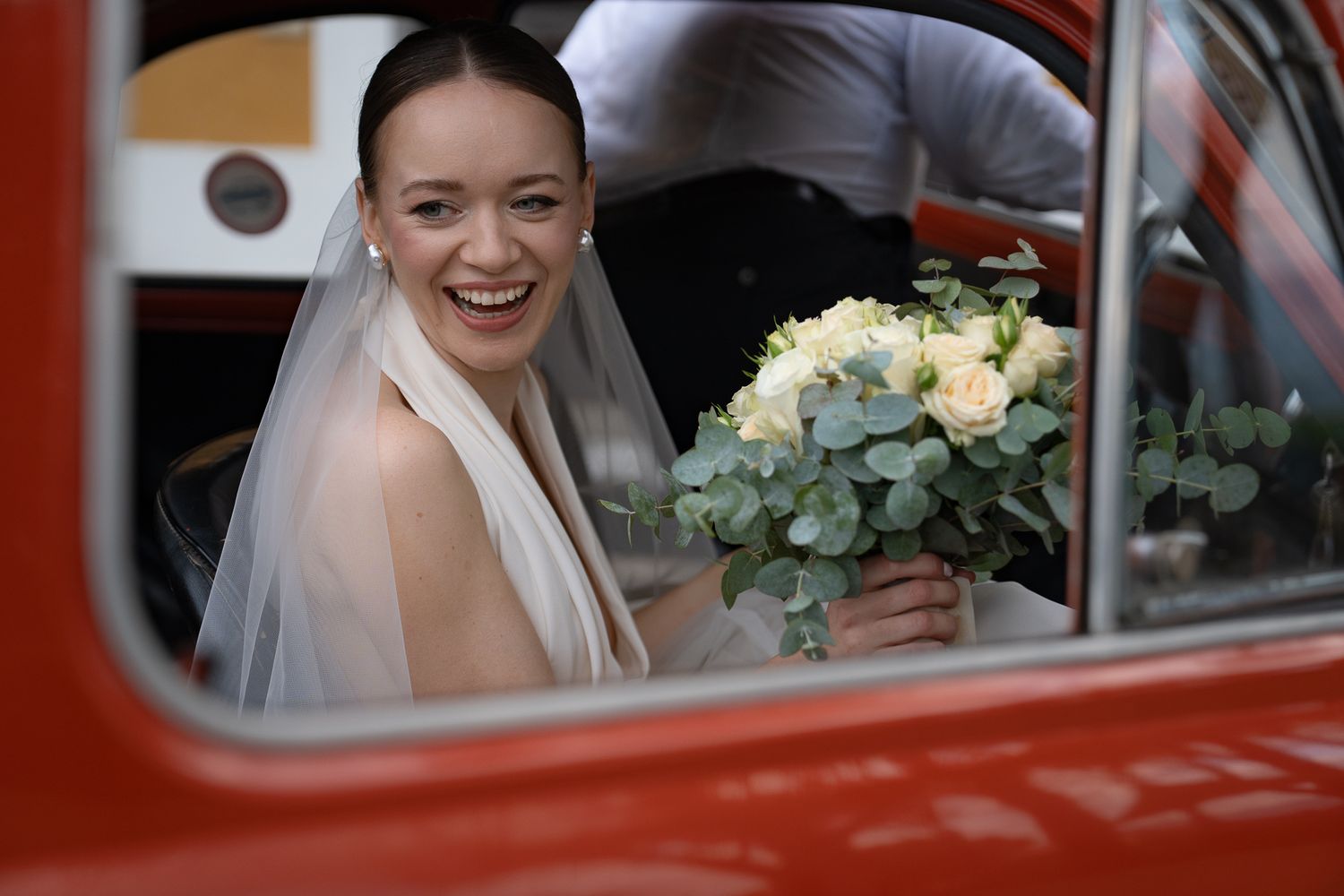 A person in a wedding dress smiles joyfully while sitting in a vintage red car.
