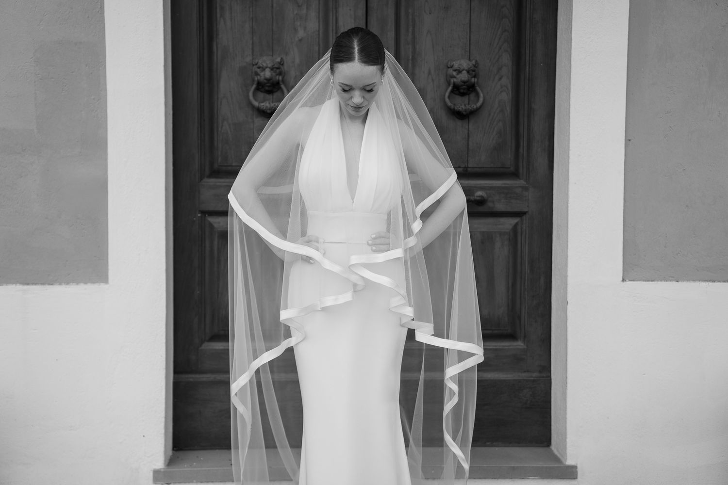 A bride in a flowing white wedding dress and veil stands in front of a white building entrance with wooden doors.
