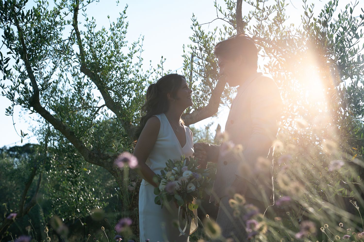 Couple sharing a romantic moment in a sunlit garden, bride holding a bouquet, golden hour light filtering through olive trees in Umbria.
