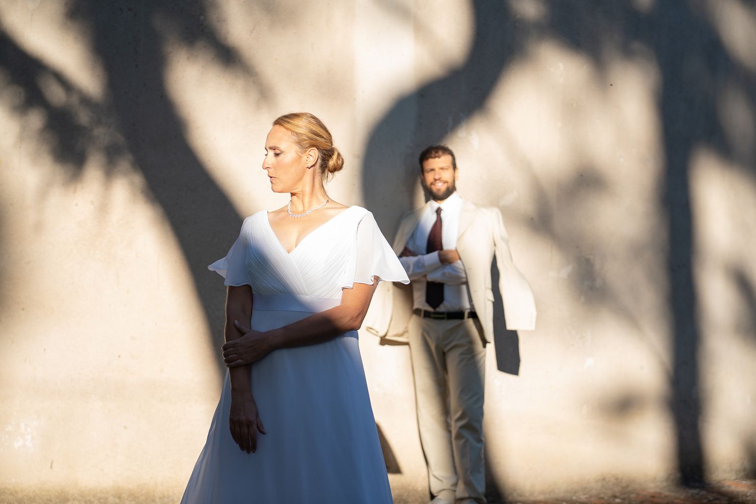 A bride and groom pose against a white wall with dramatic palm tree shadow patterns during their wedding against a church in Montone, Umbria