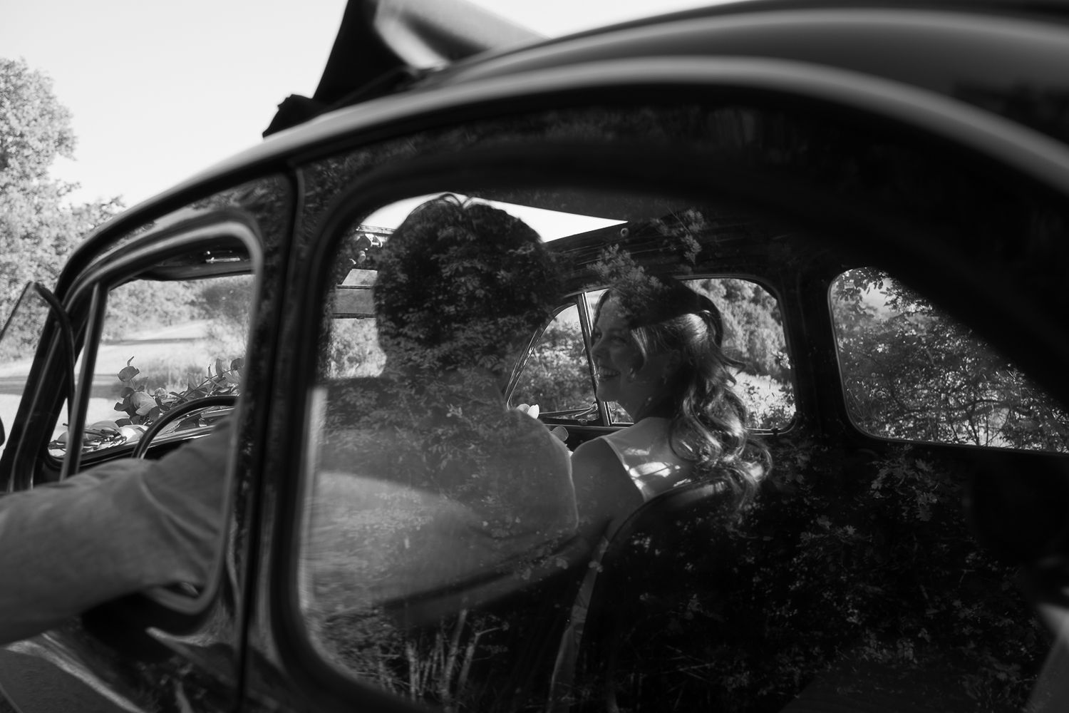 Black and white photo of a couple kissing inside a vintage car, with trees reflected in the window.