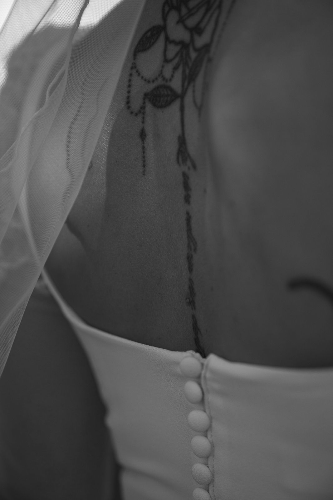 A black and white close-up photograph shows the back of a dress with delicate button details and necklace.