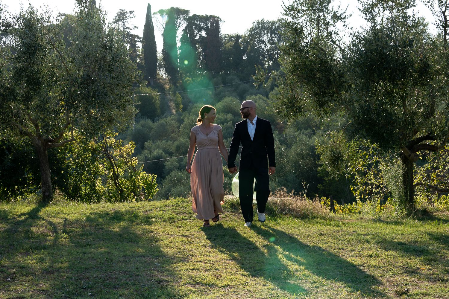 A couple walking hand in hand through a sunlit olive grove in the countryside.