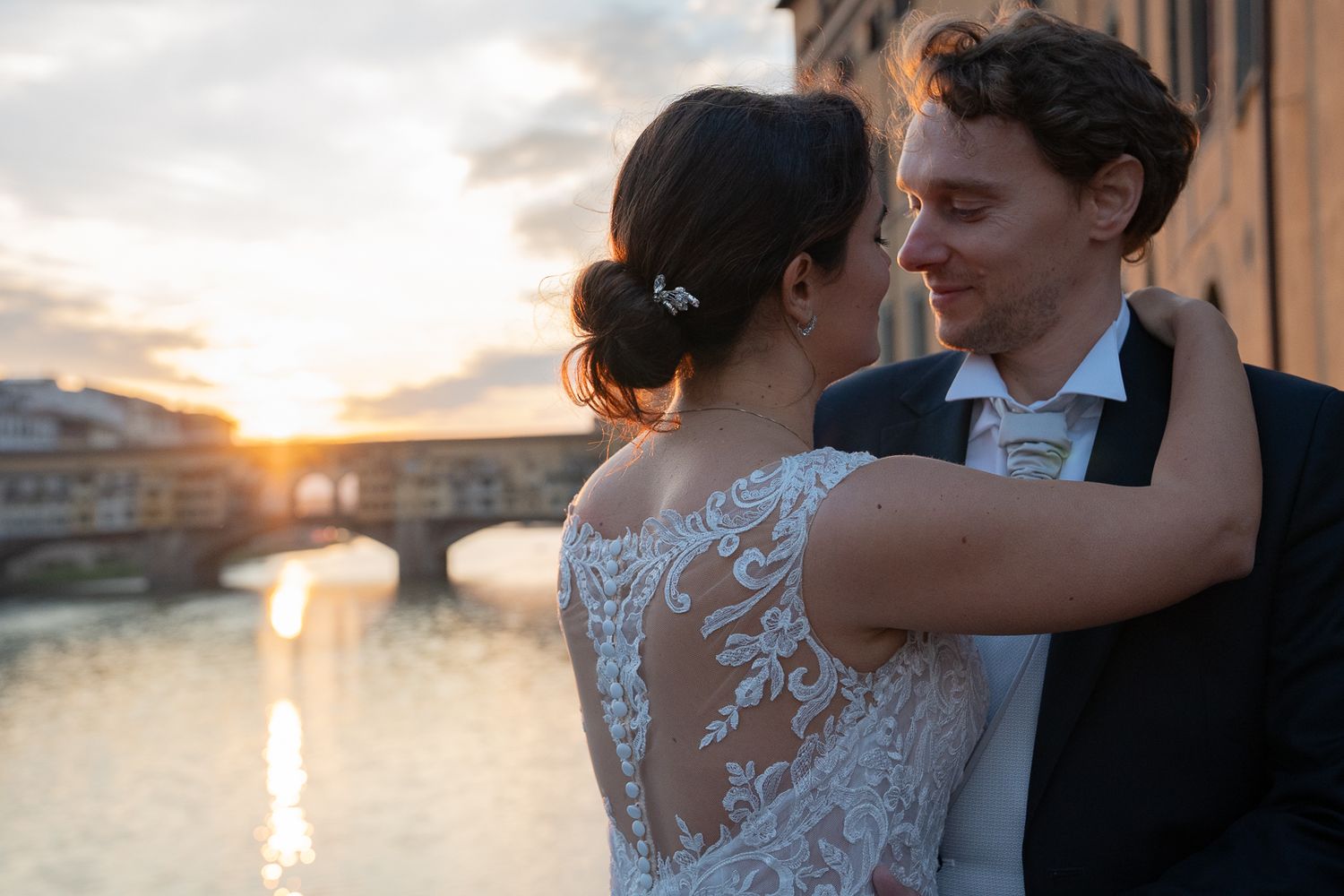 Romantic couple in wedding attire embrace while dancing at sunset near a waterfront.