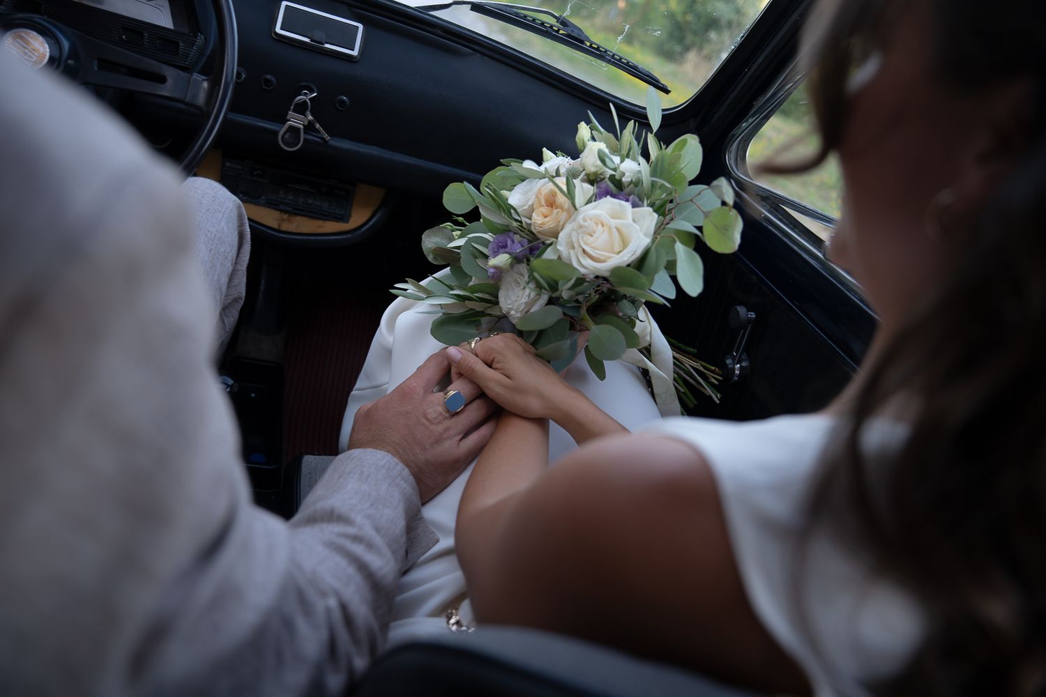 Newlywed couple holding hands with a white floral bouquet inside a vintage car Fiat 500 on their wedding day in Umbria, Italy