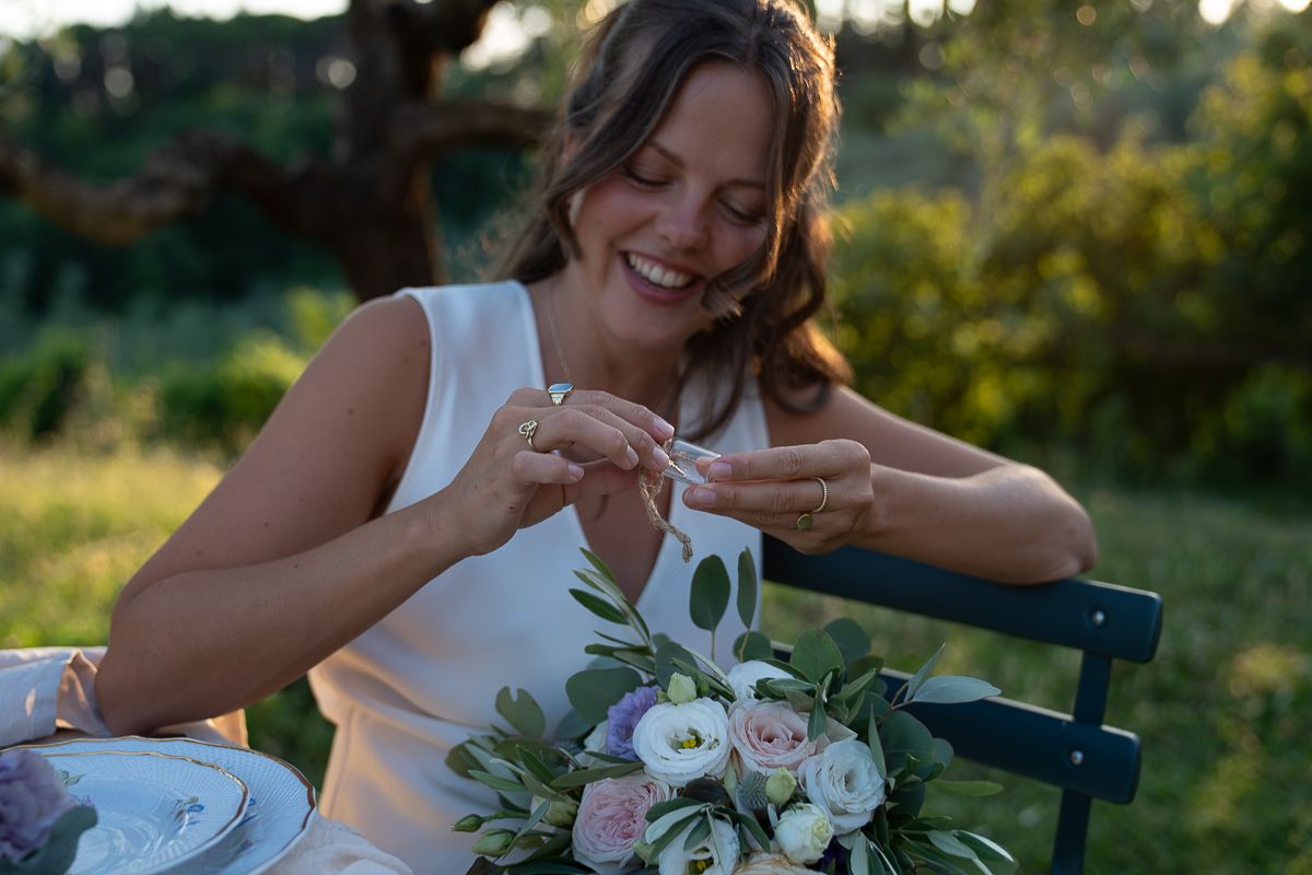 Smiling bride in white dress arranging flowers at outdoor table with bouquet of pink and white roses at golden hour at Casale San Cristoforo