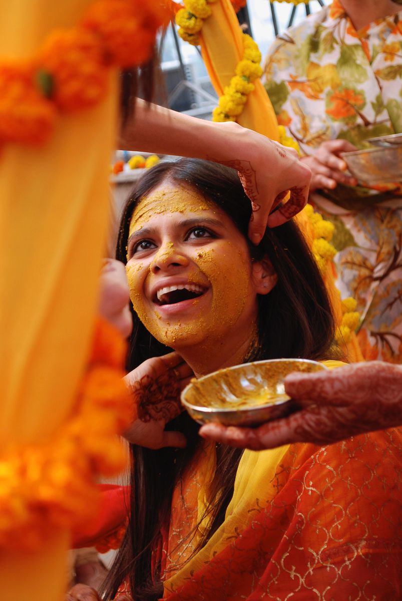 Smiling bride during Haldi ceremony with turmeric paste applied on face, surrounded by marigold flowers.