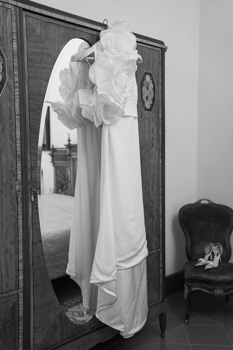 Black and white photo of an elegant white wedding dress with ruffled top hanging on a vintage wooden wardrobe mirror.