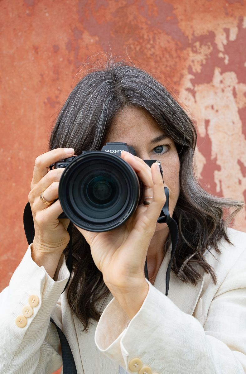 A photographer in a white sweater demonstrates three angles of holding a camera against a rustic orange wall background.