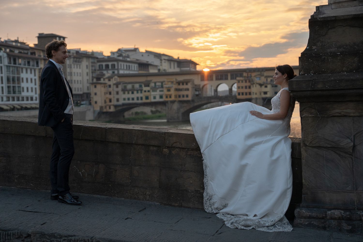 A person in a flowing white dress stands on the Ponte Santa Trinita bridge in Florence at sunset.
