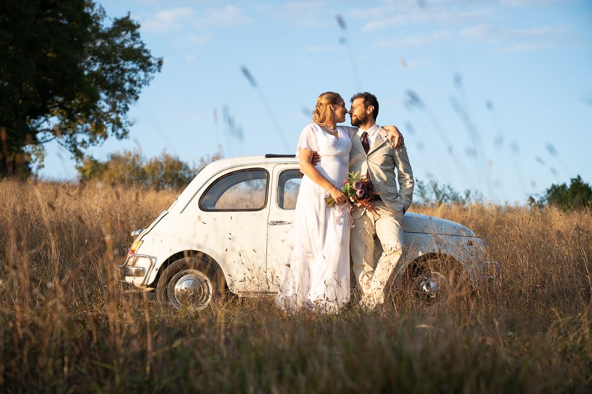 Bride and groom kissing beside a vintage white Fiat 500 in a golden field on their wedding day.