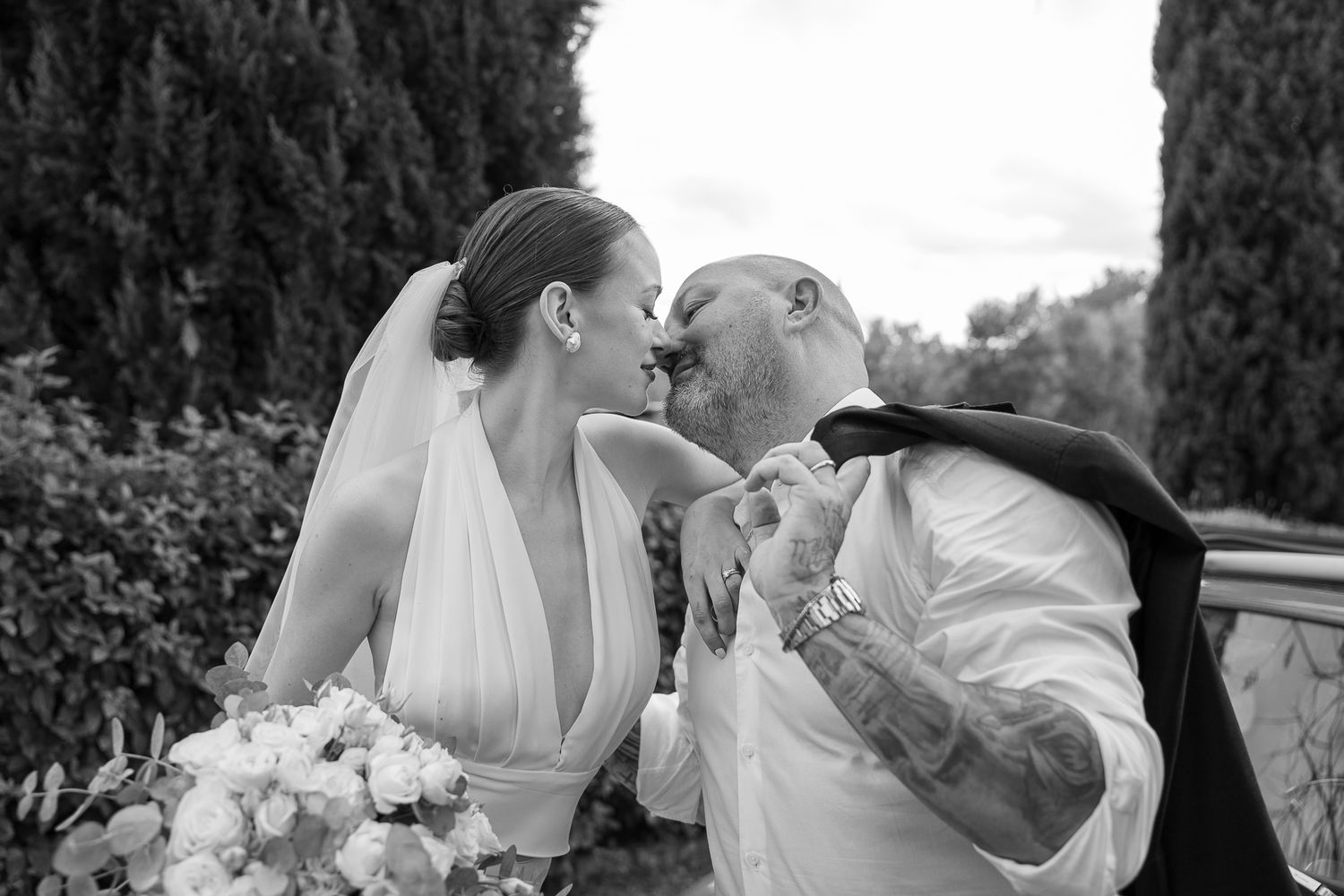 A romantic wedding couple shares tender kisses in a garden setting with cypress trees in the background.